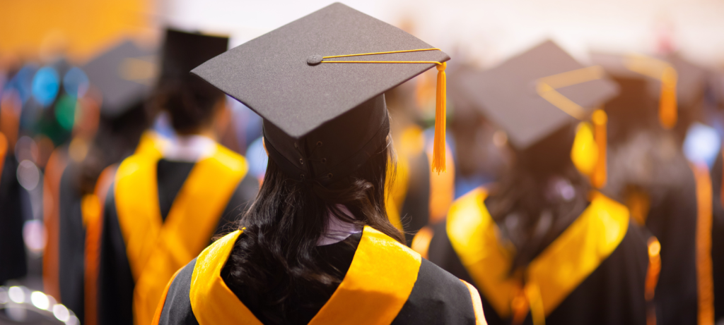 A graduate in a black cap and gold stole, seen from behind among fellow graduates at commencement